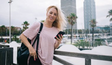 Modern cep telefonu aygıtını elinde tutan beyaz tenli çekici bir kullanıcının yarım boy portresi, akıllı telefon cihazıyla 4G 'ye bağlı turistik sırt çantalı güzel bir bayan gezgin.