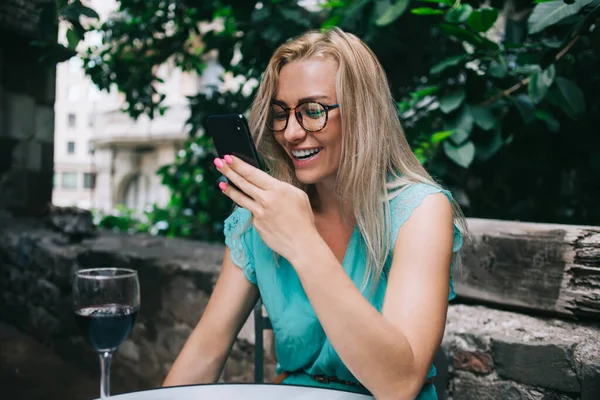 Cheerful female user in optical spectacles watching online video and laughing during wine time in street cafe, joyful Caucasian woman using cellphone device for web blogging during leisure time
