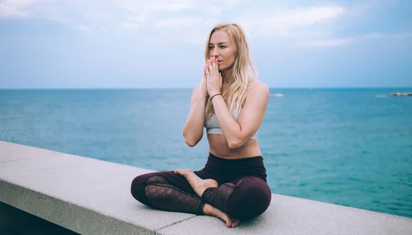 Young blond female in sports outfit sitting on embankment of sea in lotus pose and meditating looking away against overcast sky