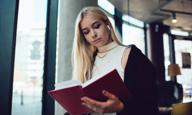 Düşük açılı, sarı saçlı, roman okuyan ve lobide durup kablosuz kulaklıkla müzik dinleyen genç bir kadın.