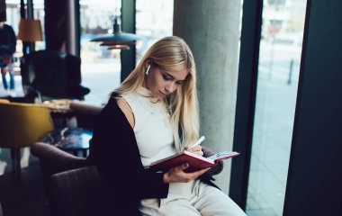 Düşünceli genç sarışın kadın kitap üzerinde çalışıyor ve kafedeki koltukta otururken kablosuz kulaklıkla müzik dinliyor.