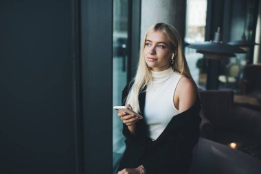 Zarif zarif bir bayan, ceketi açık, kulaklıkla müzik dinlerken kafenin kapısına yakın bir yerde akıllı telefon kullanıyor.