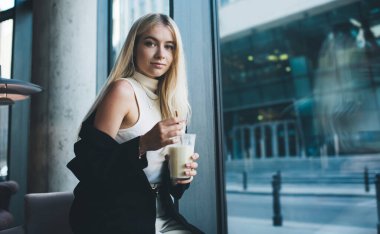 Gündüz vakti çağdaş kahve dükkanında kahve içerken çıplak omuzlarıyla kameraya bakan modaya uygun giyinmiş genç bir kadın.