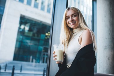 Çağdaş restoranda kahve içerken kameraya gülümseyen çıplak omuzlu, şık takım elbiseli çekici bir kadından.