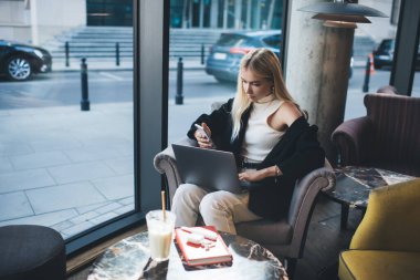 Şehir kafesinde kahve molası sırasında şık kıyafetler giyen, dizüstü bilgisayar kullanan ve akıllı telefon kullanan genç bir bayan serbest yazar.