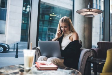 Modern kafede dizüstü bilgisayarla koltukta oturmuş telefon görüşmeleri yapan şık kıyafetli genç bir kadın serbest yazar.