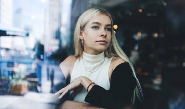 Moda kıyafetleri ve aksesuarlarıyla modern kafede oturan düşünceli sarışın kadının camdan manzarası ve gün ışığında dalgın dalgın bakışları.