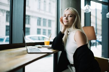 Sarışın, genç bir bayan, elinde bir bardak içki ve kafede dizüstü bilgisayarla kameraya bakıyor.