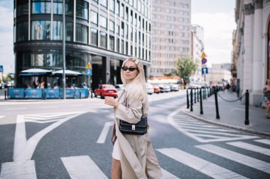 Paltolu ve güneş gözlüklü şık genç bir bayan yaya geçidinde durup yolda yürüyor ve omzunun üstünden bakıyor.