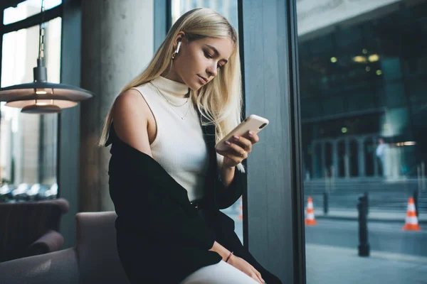 都会のロビーで椅子の側に座っている間 無線イヤフォンで携帯電話でビデオを見て流行の服で深刻な女性のフリーランサー — ストック写真