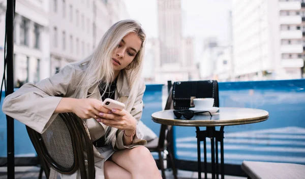ダウンタウンの昼間にストリートカフェでコーヒーを楽しみながら 流行の服の閲覧スマートフォンで現代的な女性に焦点を当て 画面を見て — ストック写真