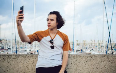 Yakışıklı, kıvırcık saçlı, sıradan tişörtlü, kayalık çitlere yaslanan ve Barcelona 'nın bulanık arka planında renkli koyu olan cep telefonuyla selfie çeken adam.