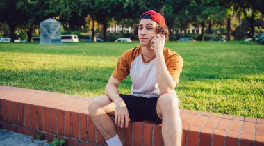 Genç, kıvırcık saçlı, sıradan giysili, kırmızı şapkalı, çitte oturan ve Barcelona 'da cep telefonuyla konuşurken kameraya bakan bir adam.