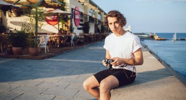 Yakışıklı, kıvırcık giyinmiş genç adam Barcelona 'nın deniz kıyısında taş bir çitin üzerinde oturmuş kameraya bakıyor.