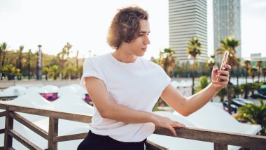 Kıvırcık sırtlı, günlük kıyafetli bir adam modern Barcelona şehrinde cep telefonuyla selfie çekiyor.