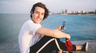 Arka plandaki berrak su ve bulutlu gökyüzü üzerinde telefon kullanarak selfie çeken genç adamın yan görüntüsü