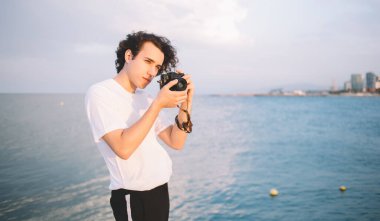 Düşüncelere dalmış gündelik giysiler içinde odaklanmış genç bir adam güneşli bir gecede deniz kenarında dikilirken fotoğraf çekmeye gidiyor.