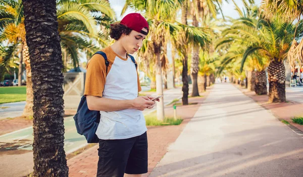 Modayı izleyen genç adam kırmızı şapkalı ve sırt çantalı ayakta duruyor ve Barcelona 'da avuç içleriyle sokakta cep telefonundan mesajlaşıyor.