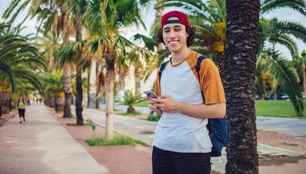 20 yaşındaki neşeli turistin yarım boy portresi güneşli Palm Caddesi 'nde duruyor ve kameraya gülümsüyor, genç milenyum kullanıcısı eğlence kaçamağı sırasında akıllı telefon cihazıyla seyahat ediyor.