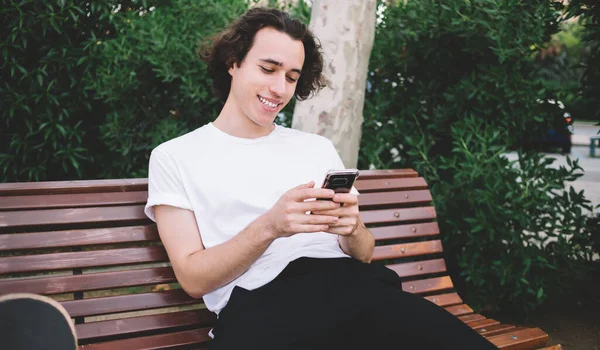 Young guy in casual clothes sitting on bench in green park and text messaging on smartphone while looking at screen and smiling