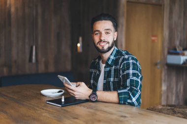 Modern kafede çalışırken akıllı telefona bakıp kahve içen ve kameraya bakan sıradan tişörtlü mutlu sakallı adam.