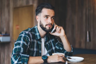 Ekose gömlekli ve kol saatli dalgın sakallı adam modern kafede kahve içerken akıllı telefondan konuşuyor ve başka tarafa bakıyor.