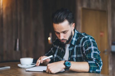Elinde ekoseli gömleği olan konsantre sakallı bir erkek kafedeki ahşap masada oturmuş kahve fincanıyla uzaktan çalışıyor.
