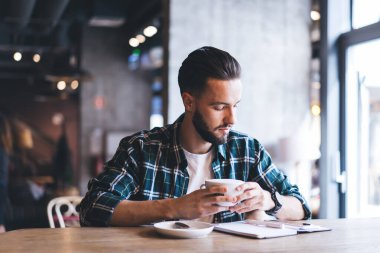 Yoğunlaşmış gömlekli erkek müşteri günlük planlayıcı okuyor ve tavan arası kafeteryasında oturup iş planları yaparken bir fincan kahve içiyor.