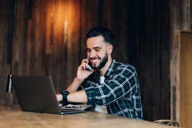 Kareli gömlekli, mutlu, serbest sakallı, cep telefonuyla konuşan, dizüstü bilgisayara bakıp gülümseyen ve karanlık kafeteryadaki ahşap iç mekânlı masada oturan erkek.