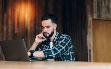 Odaklanmış, sakallı, günlük giysiler giyen ve ahşap odada çalışırken cep telefonuyla konuşan genç bir adam.