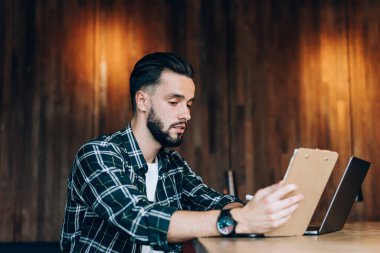 Düşünceli, serbest çalışan, günlük giyimli genç bir erkek. Yaratıcı kafe projesinde çalışırken bilgisayar ve günlük planlamacıya bakıyor.