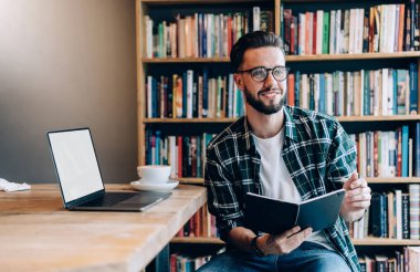 Positive young guy in glasses and trendy outfit reading book while sitting at table with laptop and cup of coffee clipart
