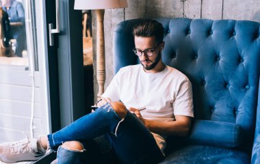 Ciddi, siyah saçlı, günlük kıyafetli, kablosuz kulaklıklı rahat koltukta oturan gün ışığında bacak bacak bacak üstüne atıp not yazan bir adam.