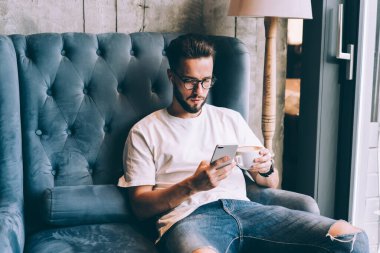 Beyaz tişörtlü, kot pantolonlu, sakallı bir adam kahve içiyor ve kanepede rahat bir şekilde otururken akıllı telefondan mesaj yolluyor.