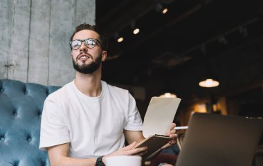 Tişört ve gözlük takan, kafedeki kanepede oturup başka tarafa bakarken panonun sayfasını çeviren genç bir adamdan.