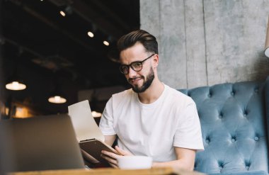 Düşük açılı mutlu sakallı adam günlük kıyafet ve gözlük takıyor. Sayfanın kapağını çeviriyor ve laptopta gülümsüyor ve kafede kanepede oturuyor.