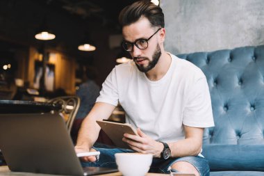 Düşük açılı, sakallı genç bir erkek elinde bir panoyla kafede oturmuş laptopta bilgi arıyor.