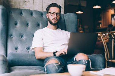 Düşünceli genç bir erkek, sakallı, günlük kıyafetler giymiş ve gözlüklü, kanepede otururken dizüstü bilgisayarında daktilo yazıyor ve başka tarafa bakıyor.