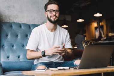 Gayri resmi kıyafetli, kahve içen ve kafeteryada dizüstü bilgisayarla kameraya bakan mutlu, genç bir erkekten.