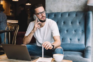 Düşünceli, sakallı, sıradan giyinmiş, gözlüklü, kafede elinde kalemle otururken cep telefonuyla konuşan ve gözlerini kaçıran genç bir erkek.