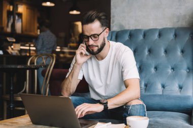 Sakallı, kot pantolon ve gözlüklü, cep telefonuyla konuşan ve kafede otururken dizüstü bilgisayarını karıştıran genç bir erkek.