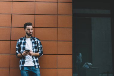 Düşünceli, traşsız, günlük giysiler içinde genç bir erkek. Şehir sokağındaki modern binanın duvarında dikilirken başka tarafa bakıyor ve akıllı telefon kullanıyor.