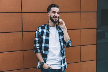 Neşeli, traşsız, günlük kıyafetlerle gezen, cep telefonuyla konuşan, şehir caddesindeki çağdaş binanın duvarının yanında duran genç bir erkek.