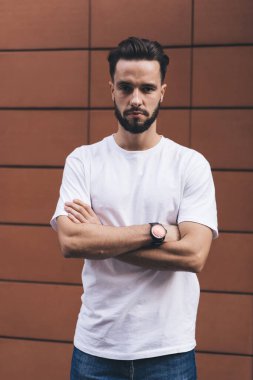 Düşünceli, genç, sakallı bir erkek, kolları bağlı, modern şehir binasının duvarında dikilirken kameraya bakıyor.