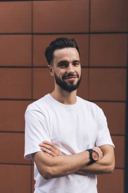 Beyaz tişört giyen neşeli sakallı hippi erkek duvarın yanında durup kameraya bakarken gülümsüyor ve dinleniyor.