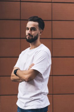 Kendine güvenen genç sakallı bir adam, sıradan kıyafetler giyip sokakta yürürken kolları çapraz bir şekilde duvara bakarken kameraya bakıyor.