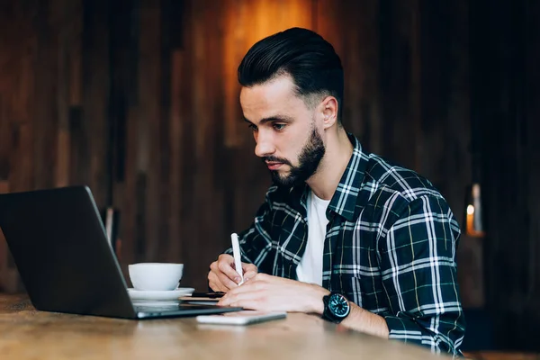 Koncentrerad Skäggig Manlig Frilansare Rutig Skjorta Tar Anteckningar Medan Sitter — Stockfoto