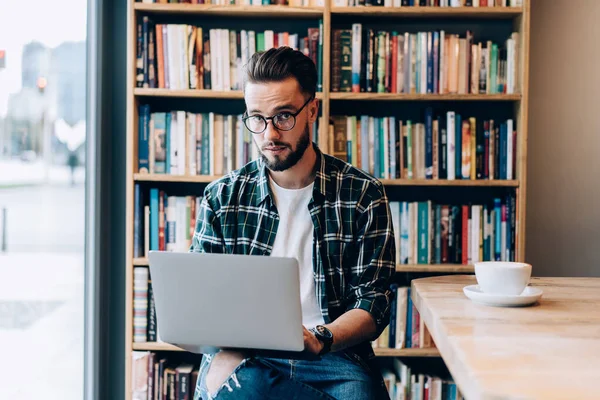 大胡子男子穿着格子衬衫 坐在舒适的工作空间里 一边浏览笔记本电脑 一边在日光下喝着大量的书和咖啡 — 图库照片