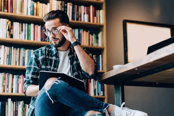 穿着手表 格子衬衫和牛仔裤的英俊大胡子男子坐在图书馆里 手里拿着黑书 翻阅着眼镜 — 图库照片
