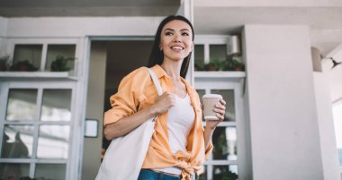 Düşük açılı, neşeli, şık, şık, esmer, günlük kıyafetler giyen, şehirde bir fincan kahveyle dikilen ve gözlerini kaçıran bir kadın.